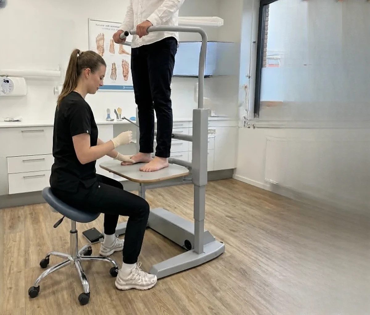 Een voetzorgspecialist zit rechtop op een kruk met de knieën onder de verhoogde voetplaat van een Orthostand en onderzoekt de belaste voet van een cliënt die op de lift staat en de handreling vasthoudt.