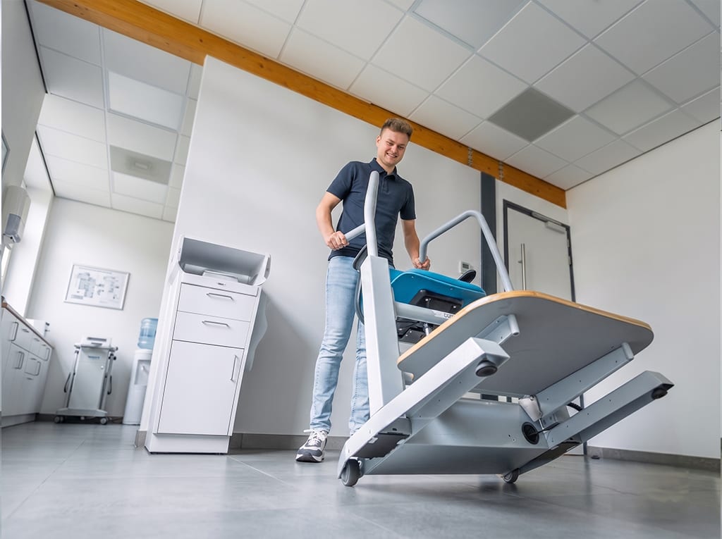 Een behandelaar rolt de Orthostand M30 op zijn wielen door een behandelkamer, met het platform gekanteld in de transportstand